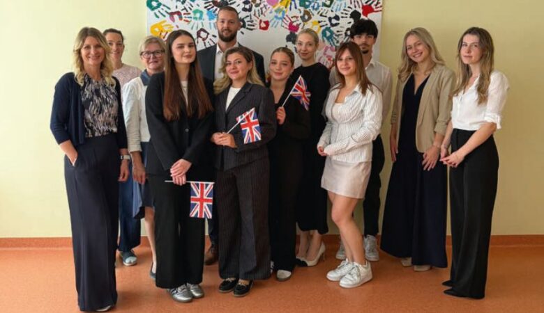 Eine Gruppe von lächelnden Studierenden und Lehrkräften steht zusammen vor einer farbenfrohen Wand mit Handabdrücken, während einige von ihnen kleine britische Flaggen halten, um das erfolgreiche Bestehen der B2-Englischprüfung zu feiern.